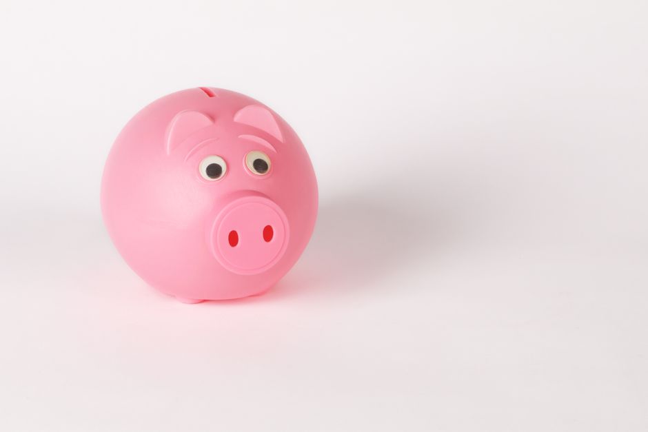 A close-up, brightly lit photo of a parent's hand and a child's hand placing a coin into a clean, white piggy bank, symbolizing saving for education.