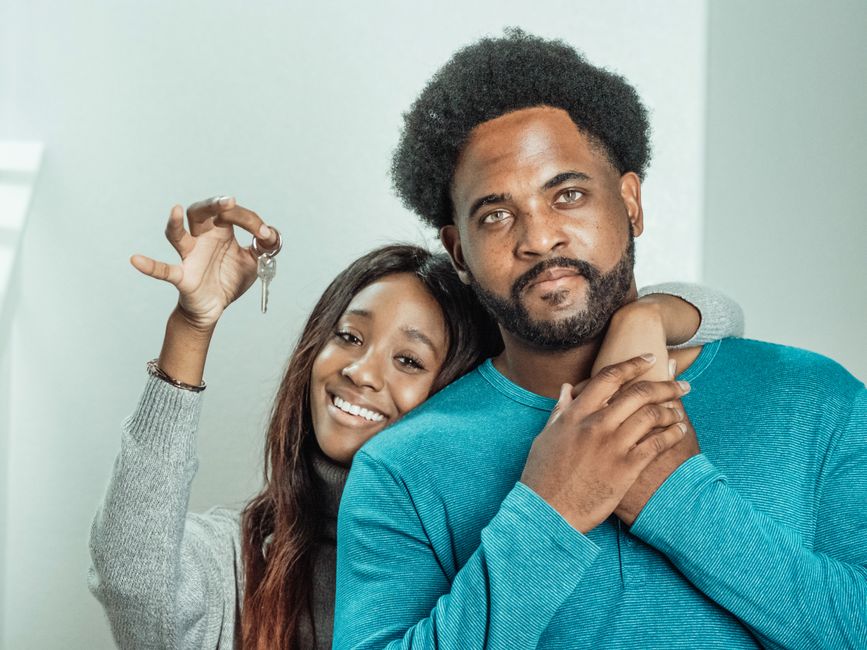 A happy couple stands inside their bright, modern home, holding a set of keys and smiling, celebrating their successful real estate transaction.